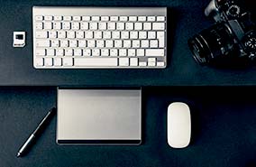 Desk with computer peripherals, including a wireless keyboard, mouse, DSLR camera, and black and silver pen